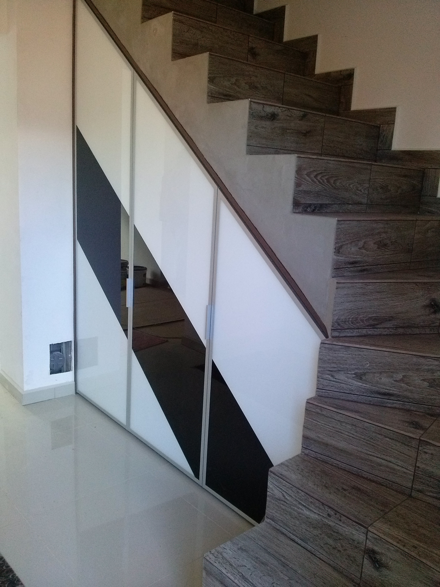 Biało-czarna szafa wnękowa pod schodami z drewnopodobnymi płytkami, widoczna instalacja elektryczna w ścianie obok.