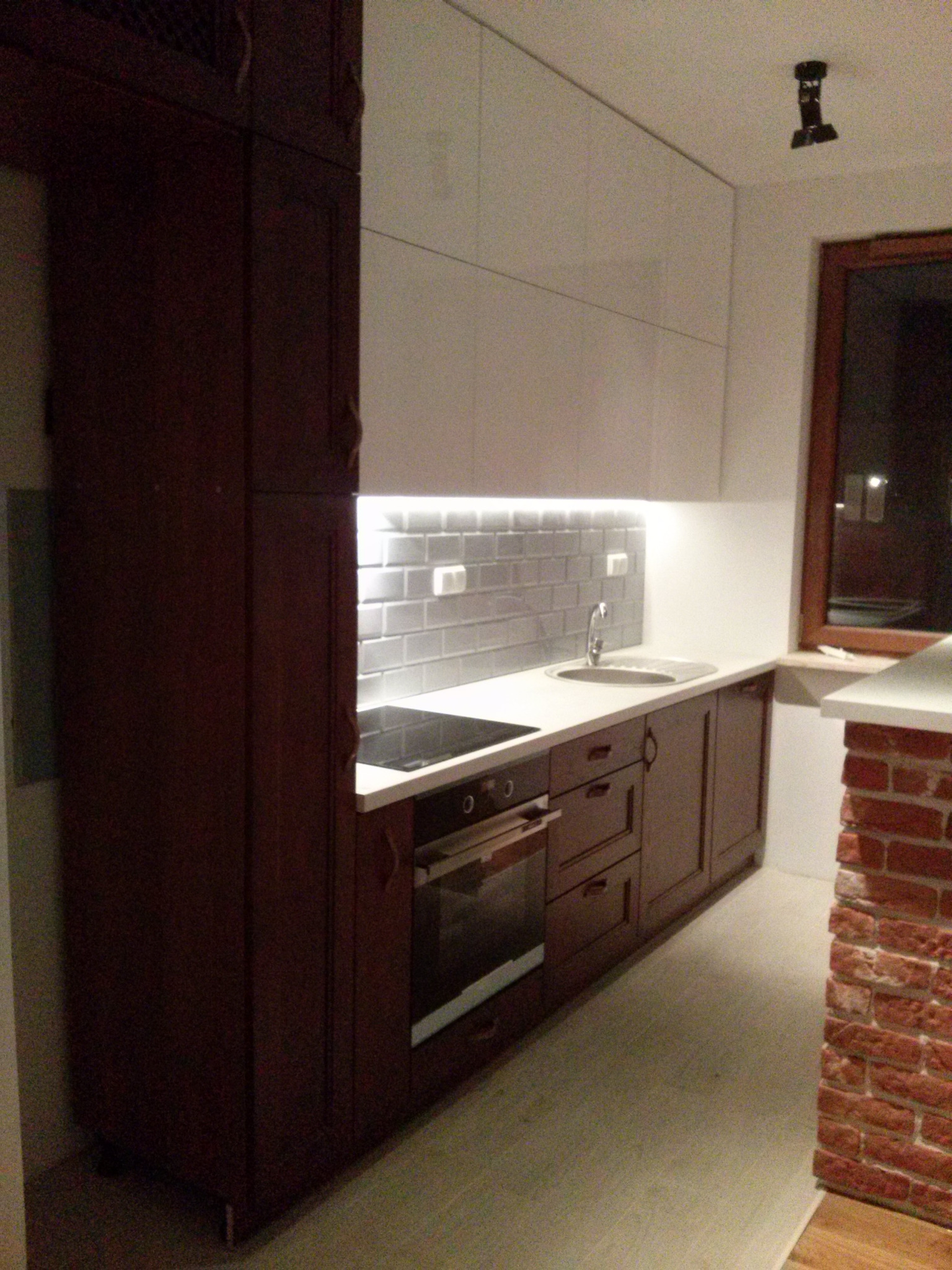 Ciemnobrązowe szafki kuchenne z białymi górnymi szafkami i szarymi płytkami na ścianie, z widoczną płytą grzewczą i piekarnikiem, oraz fragmentem ściany z cegły.