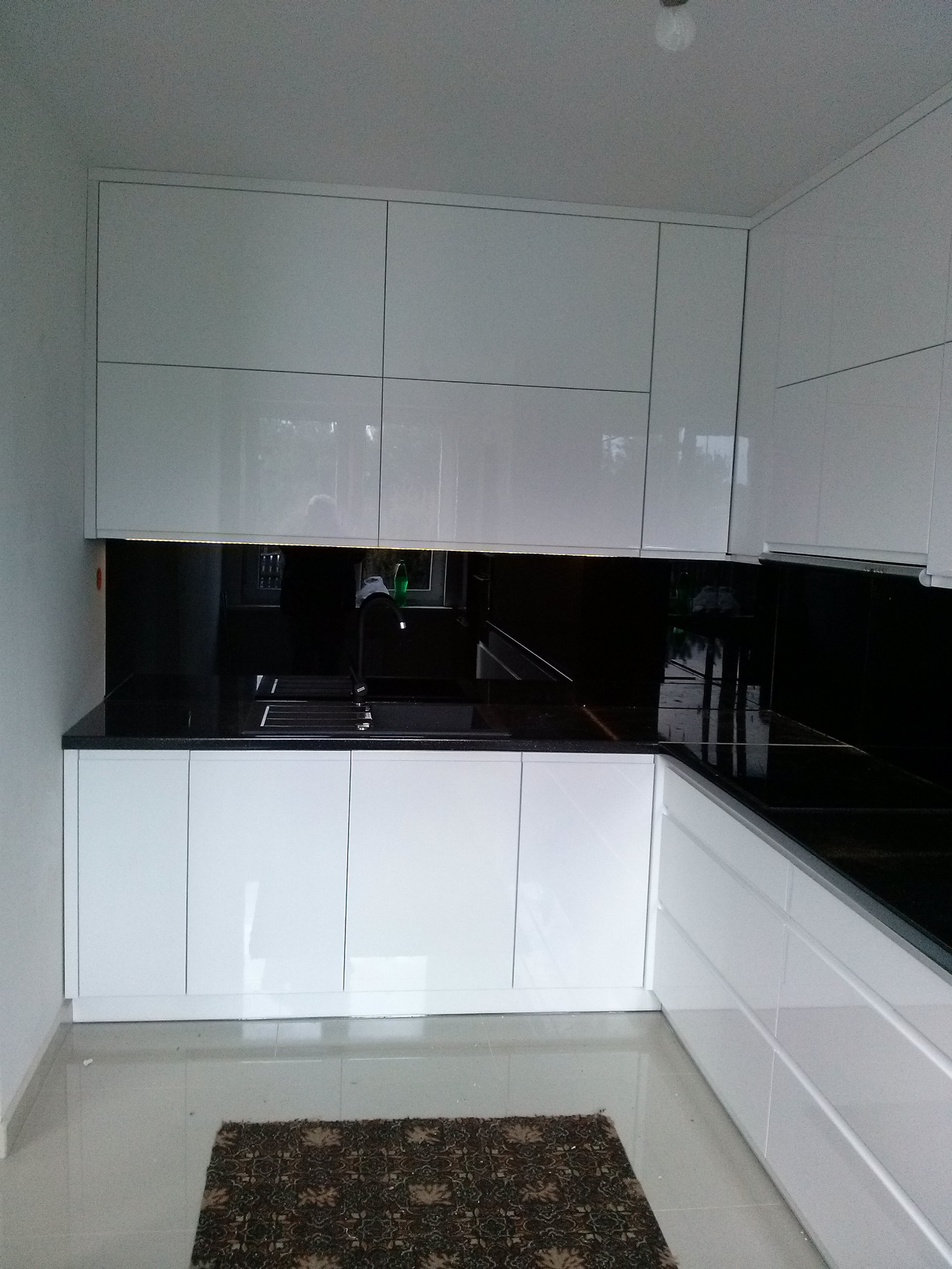 Nowoczesna kuchnia z białymi, lakierowanymi frontami, czarnym blatem i panelem nad blatem, oświetleniem LED podszafkowym i ciemnym dywanikiem na podłodze.