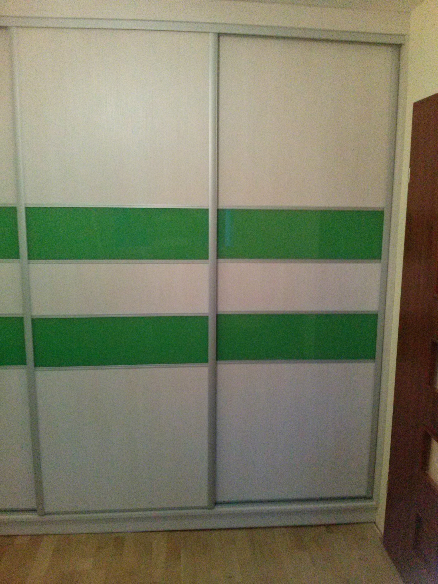 Dwudrzwiowa szafa przesuwna z białymi i zielonymi poziomymi pasami, widoczna podłoga z paneli i fragment brązowych drzwi.