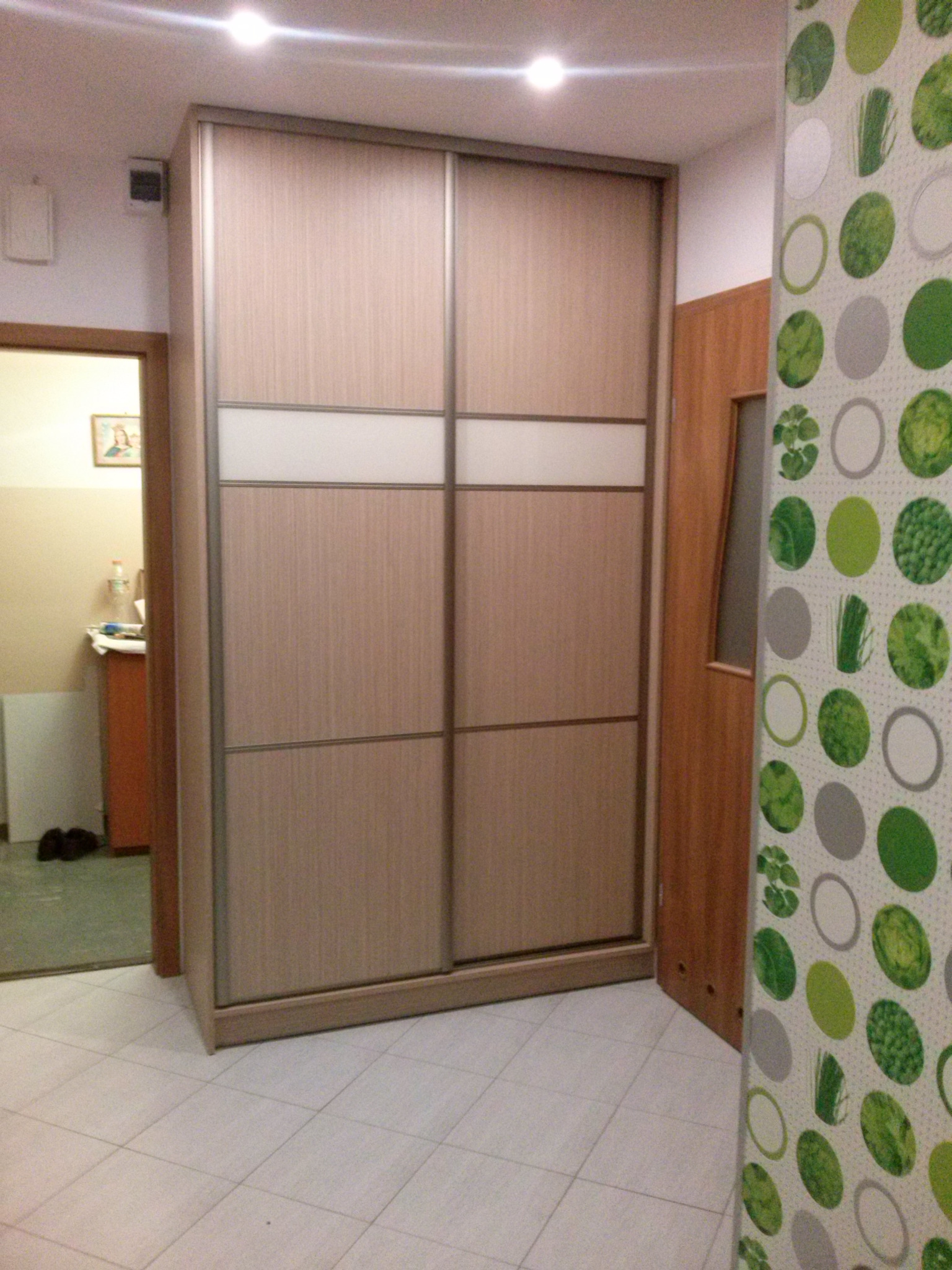 Wnętrze pokoju z szafą wnękową z przesuwnymi drzwiami w odcieniach beżu i brązu, z poziomym pasem z matowego szkła; widoczna część pokoju obok z wzorzystą tapetą.