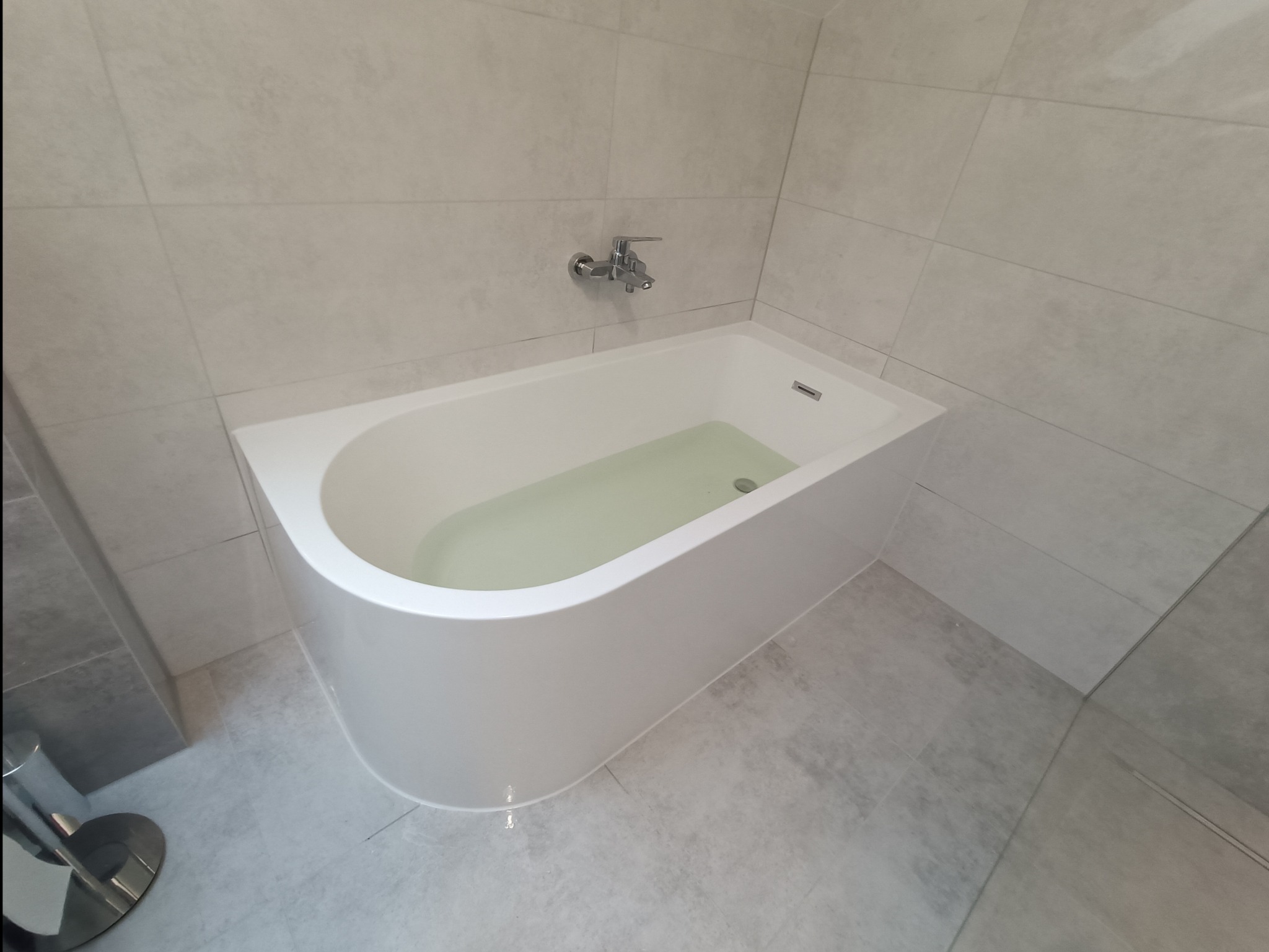 Wanna akrylowa wypełniona wodą, z chromowaną baterią ścienną, w łazience wykończonej jasnoszarymi płytkami ceramicznymi o matowej powierzchni.