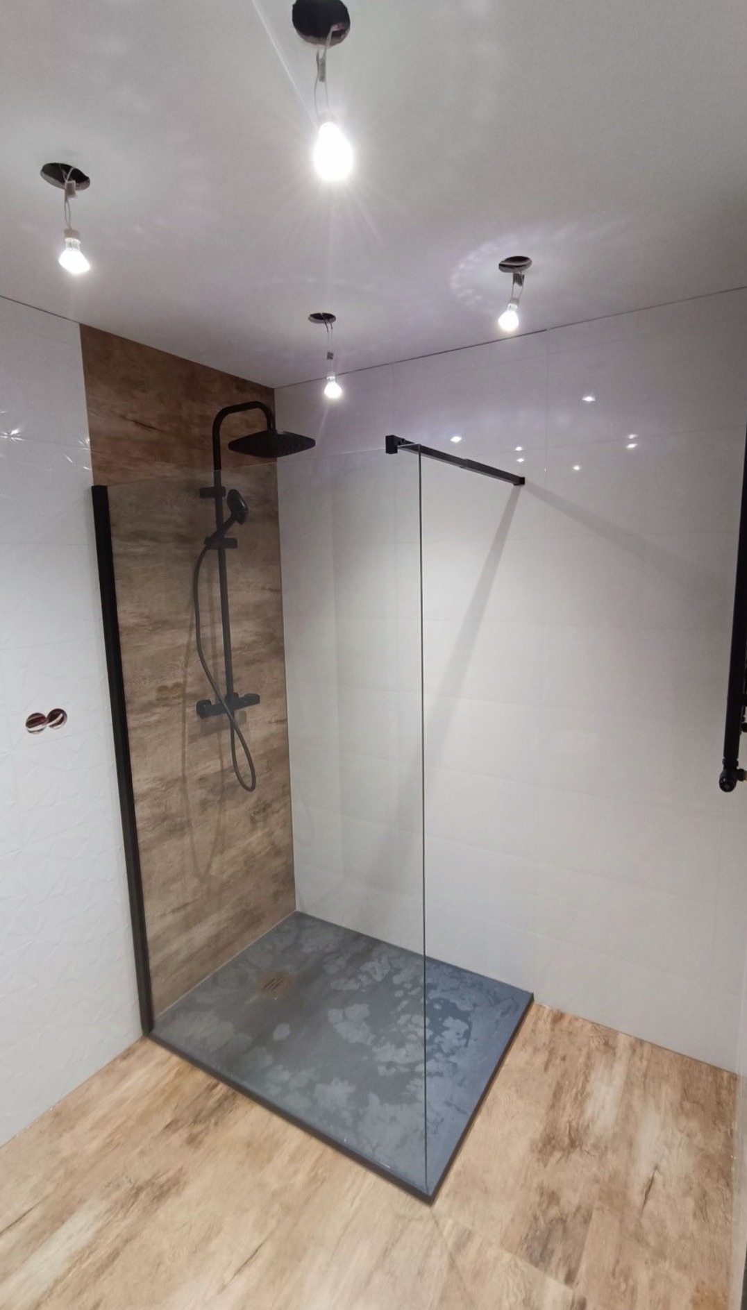 Nowoczesna kabina prysznicowa walk-in z czarną armaturą, szklaną ścianką i płytkim brodzikiem w łazience wykończonej białymi i drewnopodobnymi płytkami, z widocznymi punktowymi lampami sufitowymi.