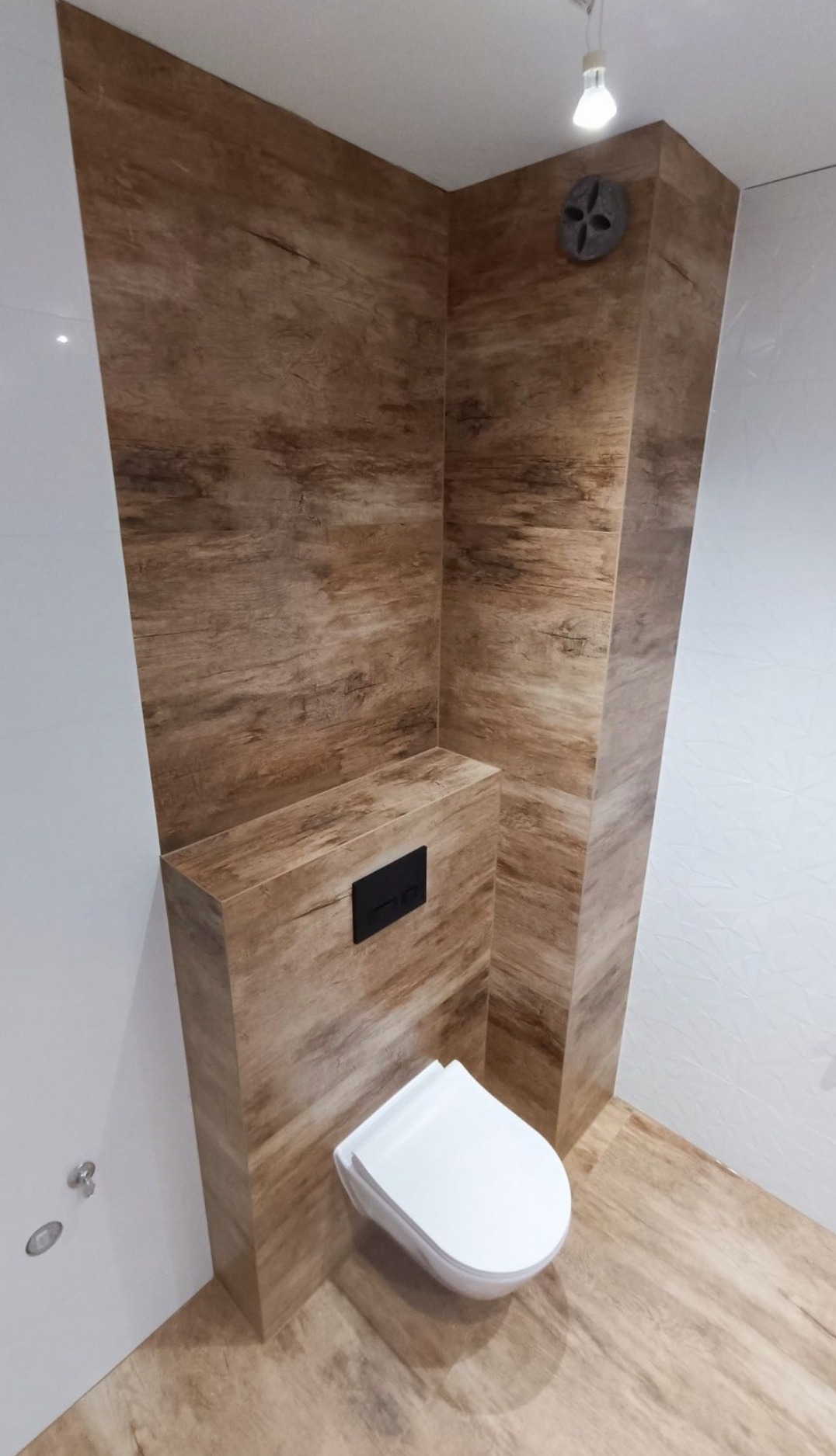 Nowoczesna łazienka z drewnopodobnymi płytkami na ścianach i podłodze, wisząca toaleta z czarnym panelem sterującym, oświetlenie punktowe, wentylator wkomponowany w narożnik.