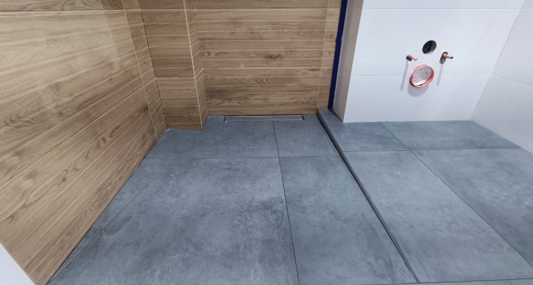 Nowoczesna kabina prysznicowa bez brodzika z odpływem liniowym, wykończona płytkami imitującymi drewno na ścianach i szarymi płytkami podłogowymi, widoczne przygotowane przyłącza wodne.