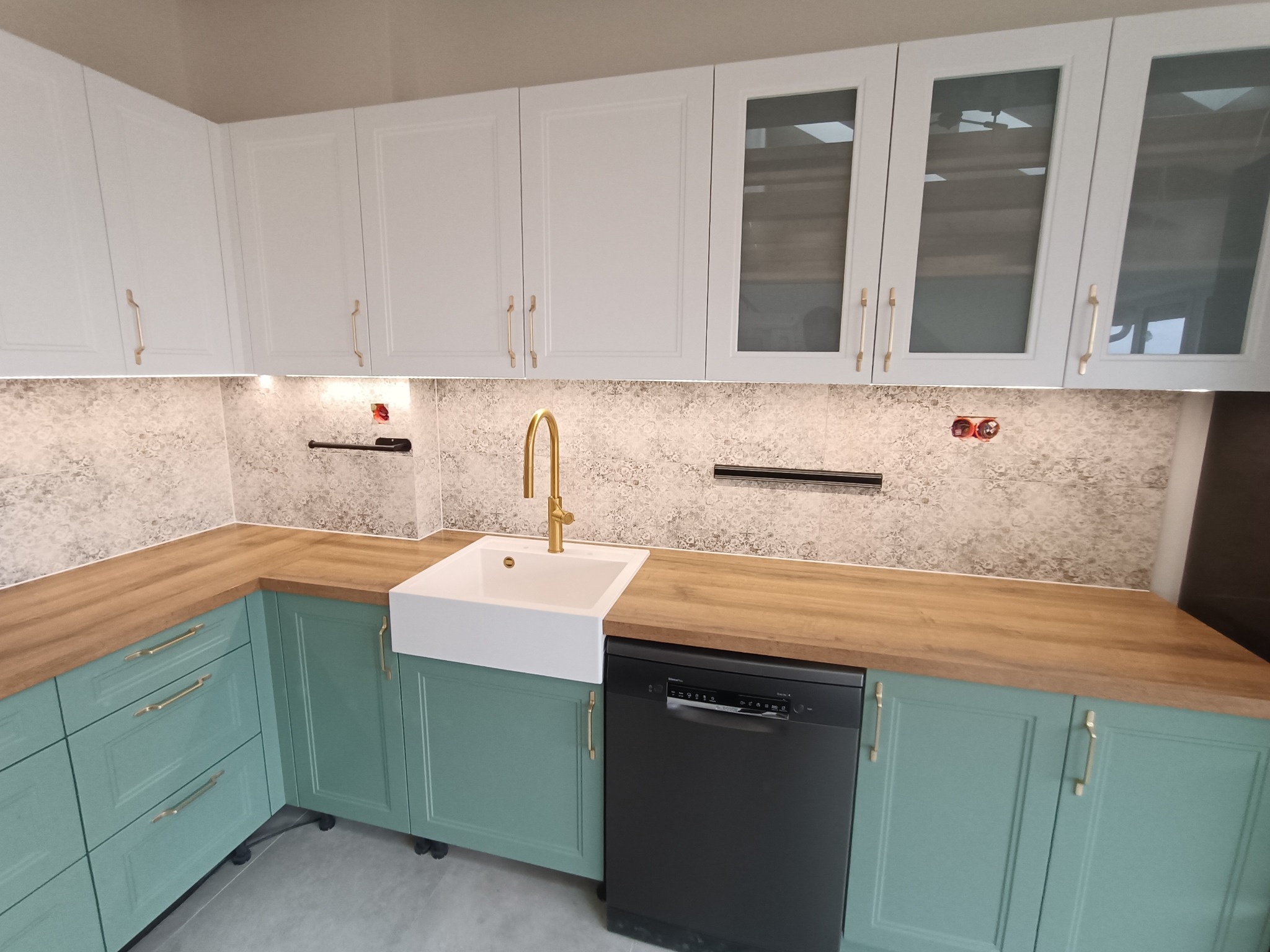 Nowoczesna kuchnia z turkusowymi szafkami, białymi szafkami wiszącymi z przeszklonymi frontami, blatem imitującym drewno i złotą armaturą, z widoczną zabudowaną zmywarką.