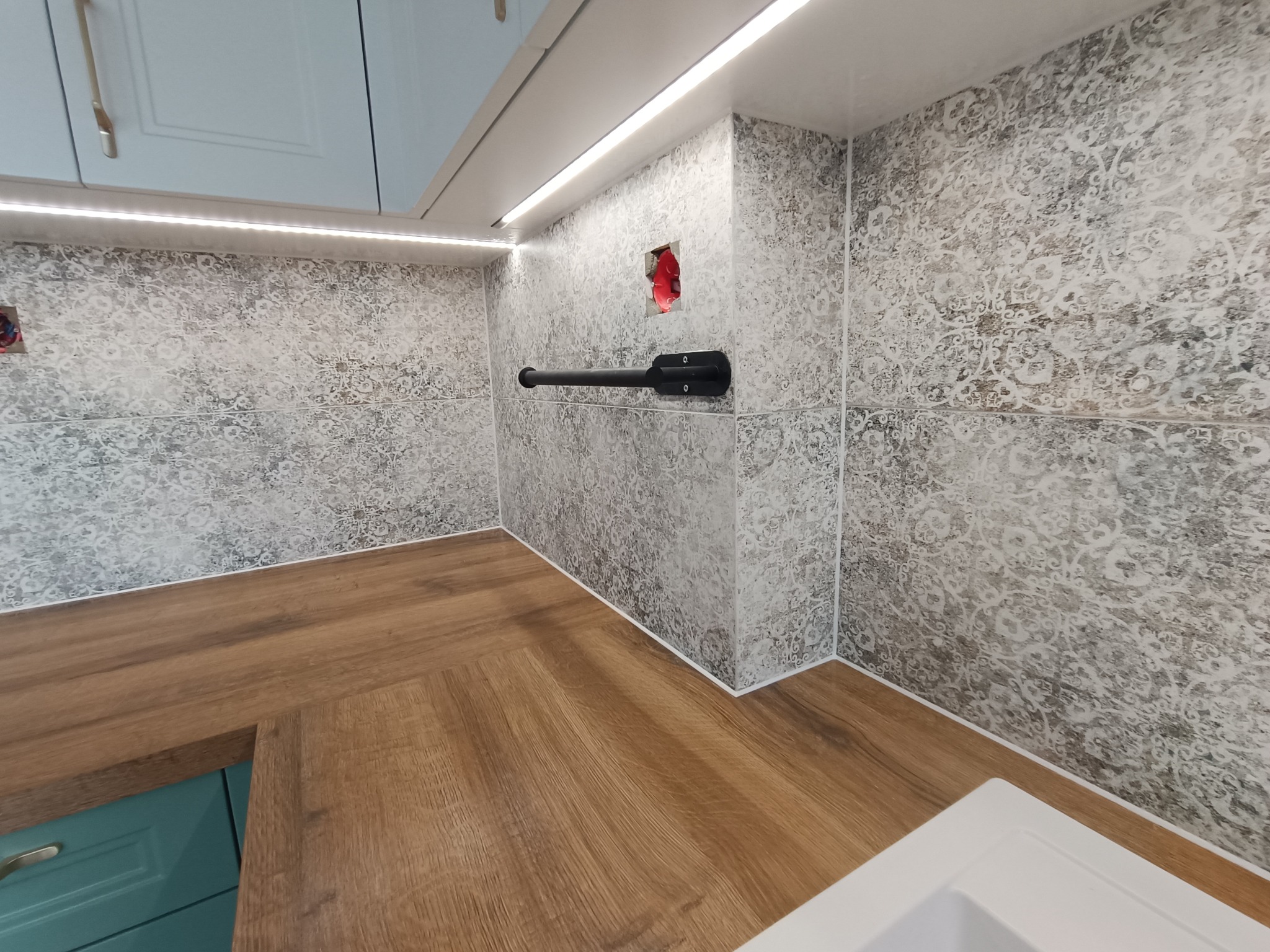 Fragment łazienki w trakcie prac wykończeniowych: ułożone płytki z wzorem, drewnopodobny blat, widoczne gniazdko elektryczne w ścianie i oświetlenie LED podszafkowe.