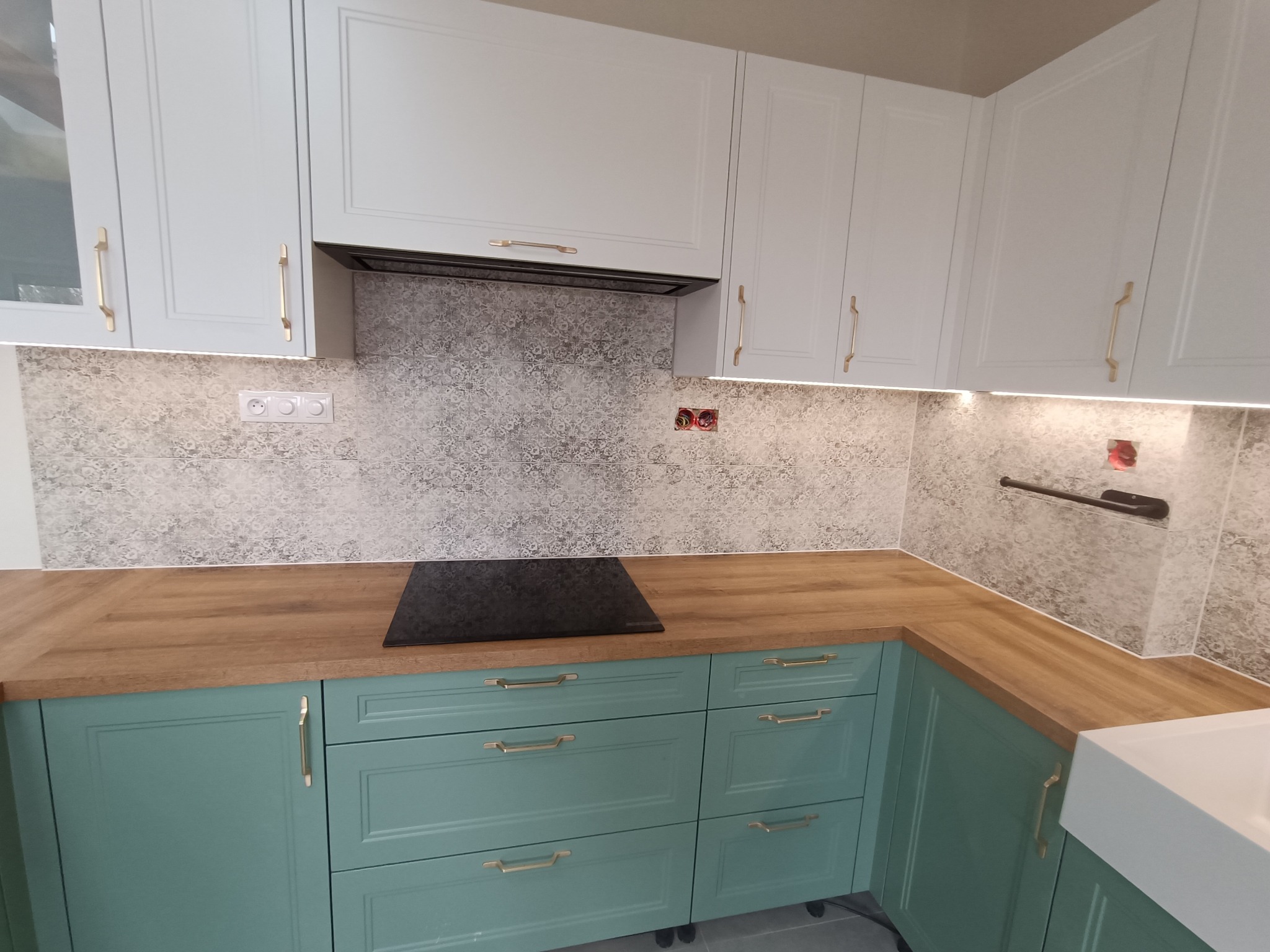 Nowoczesna kuchnia z turkusowymi szafkami dolnymi i białymi górnymi, drewniany blat, indukcyjna płyta grzewcza i dekoracyjne płytki ścienne z szarym wzorem.