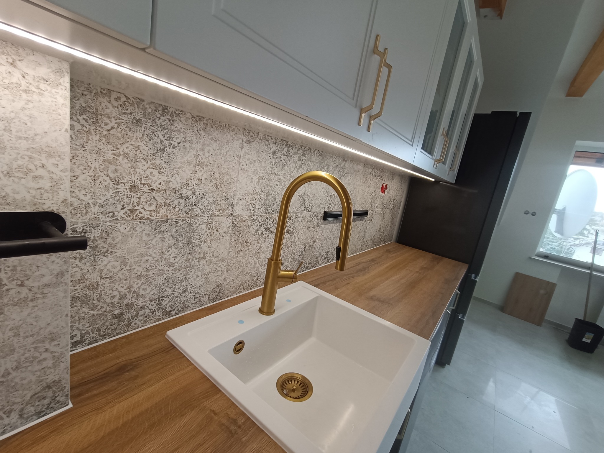 Biały zlew z złotą armaturą, zamontowany w blacie imitującym drewno, nad którym wisi szafka z złotymi uchwytami, podświetlona listwą LED, na ścianie płytki z wzorem.