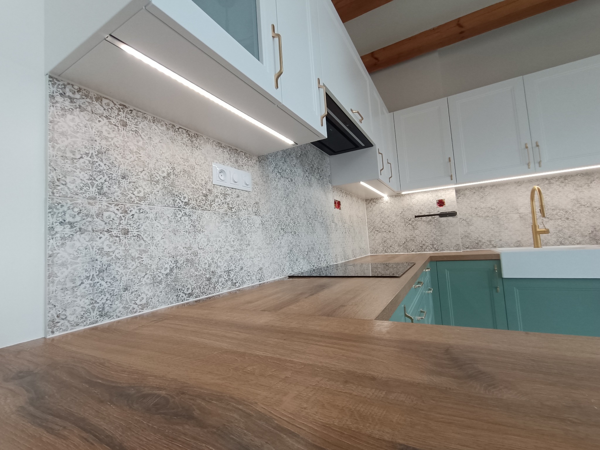 Nowoczesna kuchnia z drewnianym blatem, ozdobnymi płytkami ściennymi, białymi szafkami z złotymi uchwytami i oświetleniem LED podszafkowym.