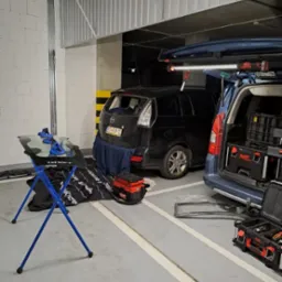 Tylna szyba samochodu Mazda 5 zdemontowana, przygotowana do wymiany w garażu podziemnym. Na stojaku leży nowa szyba z przyssawkami, obok skrzynki narzędziowe i samochód serwisowy.