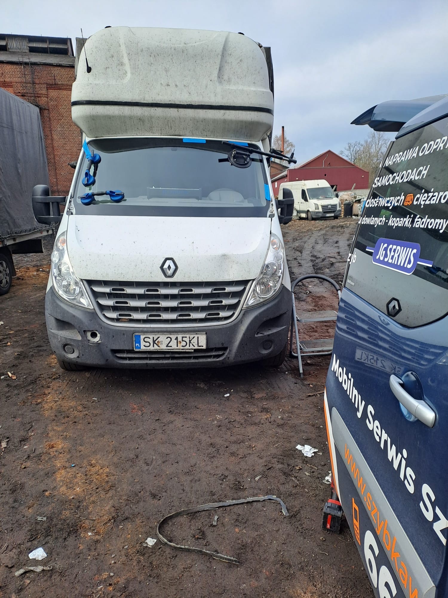 Renault Master z przyssawkami do szyb na przedniej szybie, stojący na zabłoconym placu, obok zaparkowany niebieski samochód z napisem Mobilny Serwis Szyb.