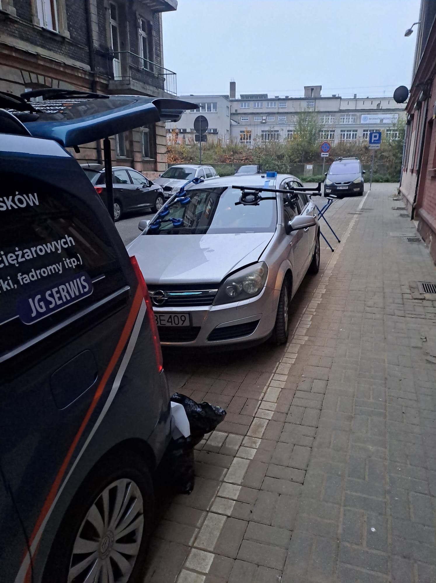 Srebrna Opel Astra z przyssawkami i uchwytami na przedniej szybie, zaparkowana na brukowanej uliczce obok ciemnego samochodu dostawczego z widocznym logo 'JG Serwis' i częściowo otwartym...