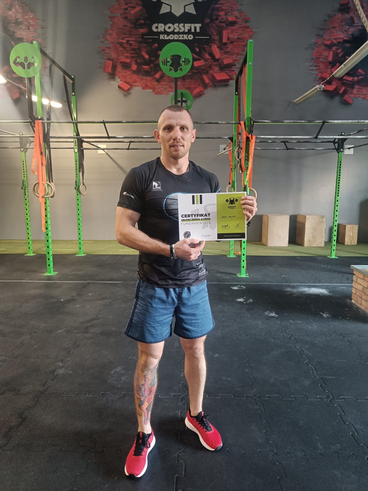 Mężczyzna w sportowym stroju trzyma certyfikat ukończenia kursu Fundamentals w siłowni CrossFit Kłodzko, z tatuażem na nodze.