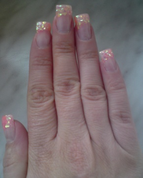 Dłoń z pomalowanymi paznokciami w odcieniach różu i brokatu, kwadratowy kształt, widoczne odrosty, marmurkowe tło.