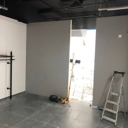 BudSeb - Wnętrze w trakcie prac: szare ściany, biała ściana z czarnym wieszakiem, drabina, podłoga z płytek, oświetlenie budowlane.