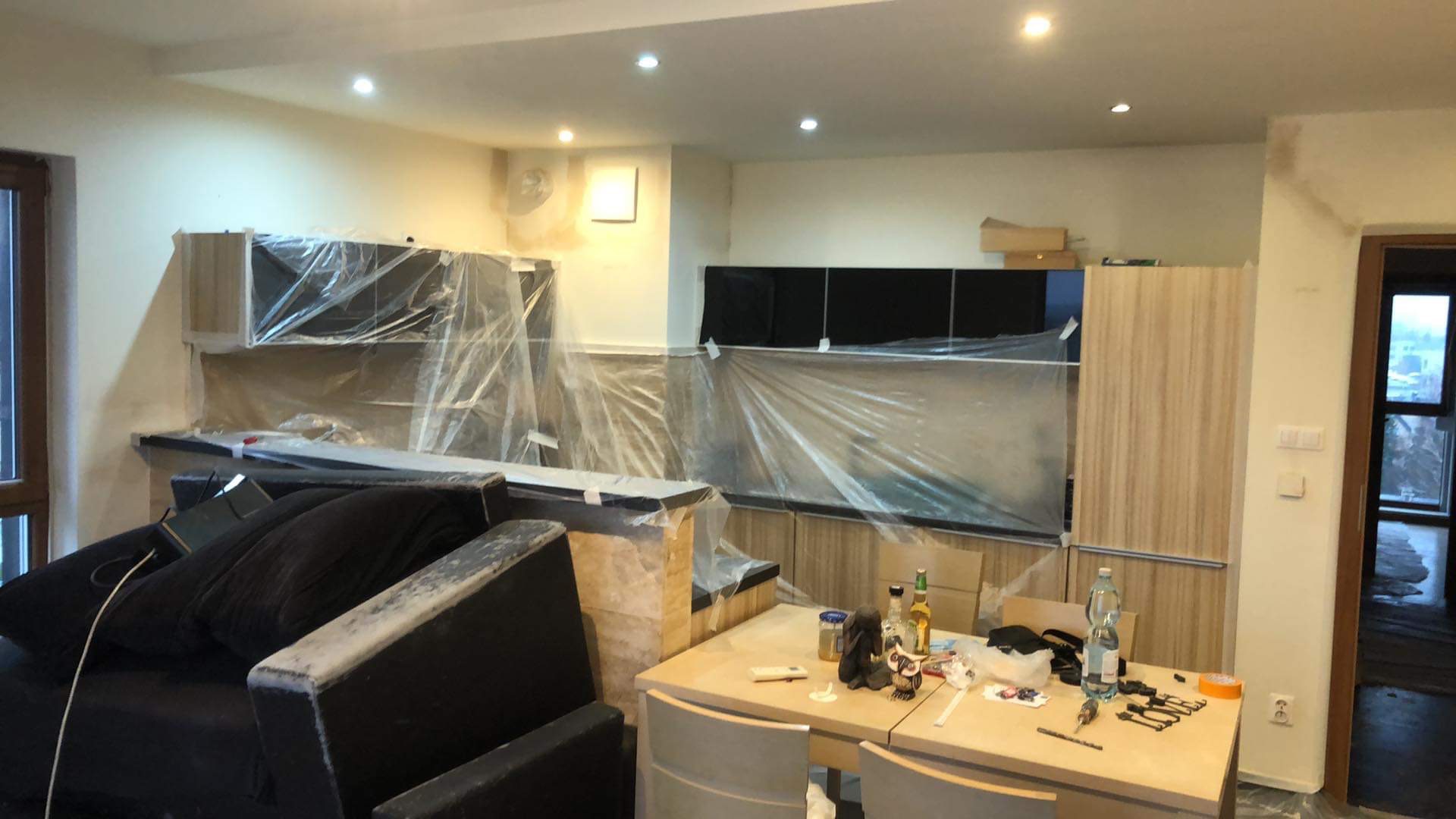 Remont pokoju: meble kuchenne zabezpieczone folią malarską, ściany przygotowane do malowania, widoczny bałagan na stole i częściowo zasłonięty mebel.