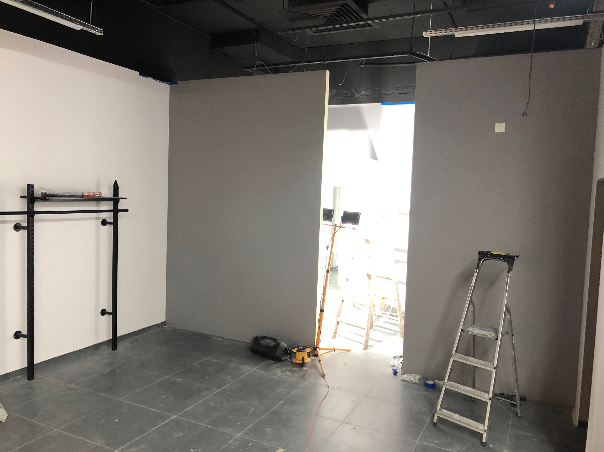 Wnętrze w trakcie prac: szare ściany, biała ściana z czarnym wieszakiem, drabina, podłoga z płytek, oświetlenie budowlane.