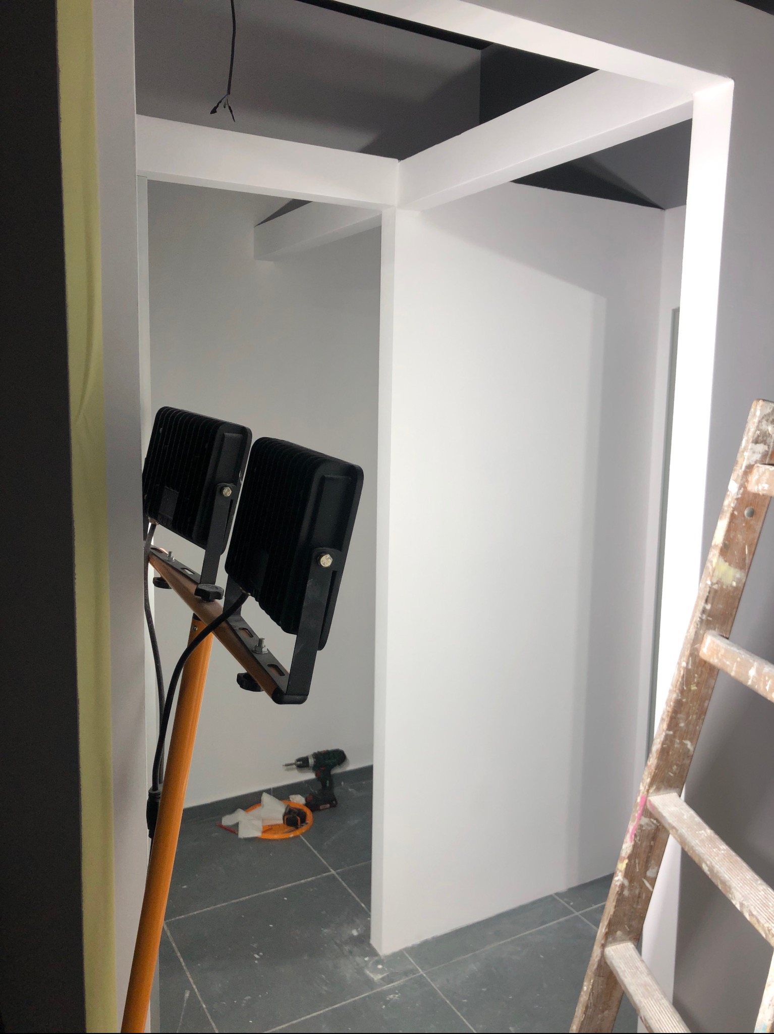 Wnętrze w trakcie odnawiania: białe ramy drzwi, szare ściany i podłoga, drabina malarska, lampa budowlana na statywie i wkrętarka na podłodze.