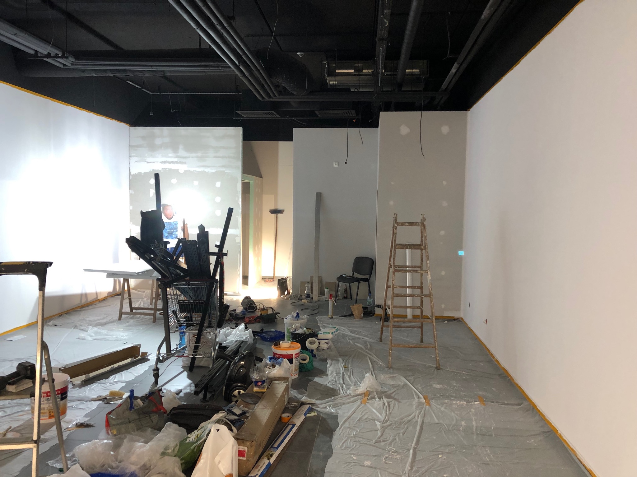 Wnętrze w trakcie prac przygotowawczych do malowania: zabezpieczona folią podłoga, drabina oparta o ścianę, widoczne puszki z farbą i narzędzia malarskie, ściany częściowo zagruntowane.