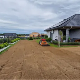 Czerwony traktor glebogryzarką przygotowuje ziemię pod trawnik na posesji z nowoczesnym domem z panelami słonecznymi.