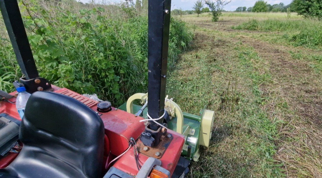 Widok z perspektywy operatora na kosiarkę trawnikową podczas pracy na polu, z widoczną częścią skoszonej trawy i wysoką trawą w tle.