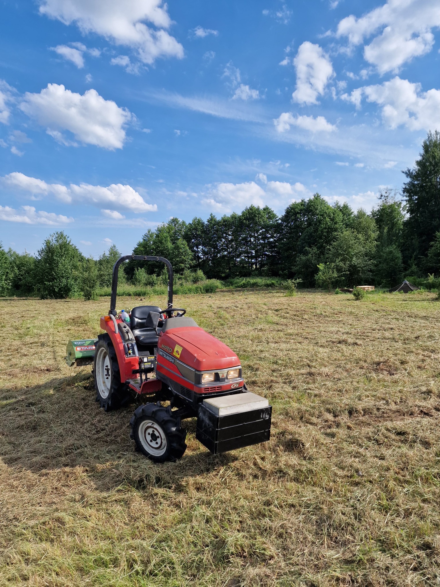 Czerwony traktor z kosiarką rotacyjną na polu świeżo skoszonej trawy, widoczne drzewa w tle i błękitne niebo z chmurami.
