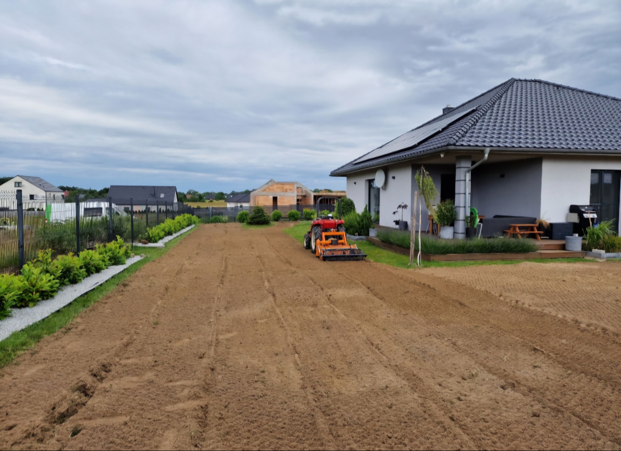 Czerwony traktor glebogryzarką przygotowuje ziemię pod trawnik na posesji z nowoczesnym domem z panelami słonecznymi.