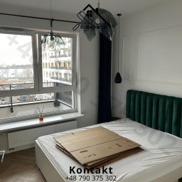 Kacper Kurkowski - Pokój z zielonym zagłówkiem łóżka, zasłonięty kartonami, widoczne dekoracyjne listwy ścienne i nowoczesne lampy wiszące.