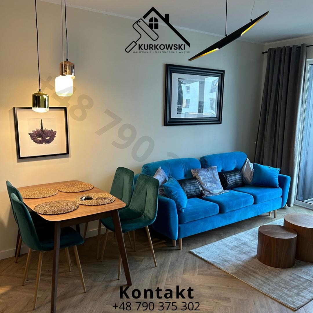Salon z niebieską sofą, drewnianym stołem jadalnym i zielonymi krzesłami, oświetlony wiszącymi lampami. Na ścianie wiszą obrazy, a na podłodze leży dywan. W tle widoczne logo firmy malarskiej.