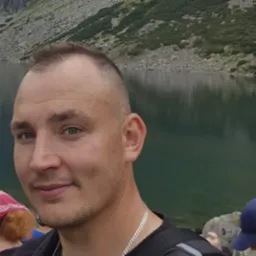 Portret mężczyzny z krótkimi włosami na tle górskiego jeziora, w tle widoczne skały i woda, z lewej strony fragment osoby z czerwoną chustą na głowie.