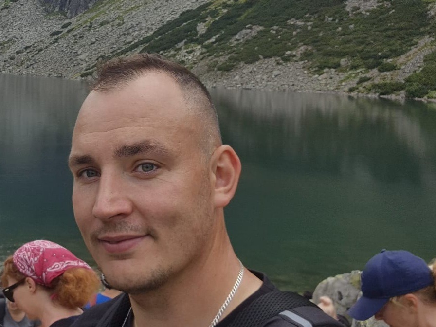 Portret mężczyzny z krótkimi włosami na tle górskiego jeziora, w tle widoczne skały i woda, z lewej strony fragment osoby z czerwoną chustą na głowie.