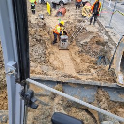 Usługi hydrauliczne wod-kan - Widok z kabiny koparki na wykop z pracownikami w żółtych kamizelkach, zagęszczających piasek ubijarką wibracyjną, z widoczną drabiną i rurami, na tle samochodu dostawczego z logo firmy.