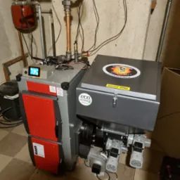 Kocioł na paliwo stałe z podajnikiem, widoczne elementy instalacji grzewczej, zbiornik wyrównawczy, sterownik z wyświetlaczem oraz naklejki informacyjne i logo producenta. Na podłodze widoczne...