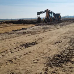 Żółta koparko-ładowarka w trakcie prac ziemnych na placu budowy, wyrzucająca urobek z łyżki, ślady opon na piaszczystym gruncie, w tle błękitne niebo.