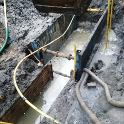 Usługi hydrauliczne wod-kan - Wykop zabezpieczony szalunkiem stalowym, wypełniony wodą i błotem, widoczne rury i węże w Wyszkowie.