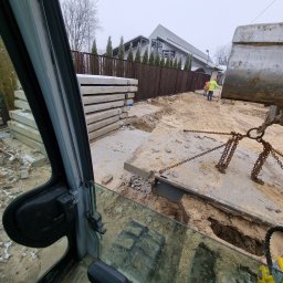 Usługi hydrauliczne wod-kan - Widok z kabiny koparki na plac budowy z betonowymi płytami i pracownikami w odblaskowych ubraniach, przygotowywany wykop pod przyłącze wodno-kanalizacyjne