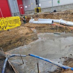 Usługi hydrauliczne wod-kan - Wykop z częściowo zalaną wodą, widoczne rury kanalizacyjne, pracownik w odblaskowej kamizelce i kasku przy wykopie, żółte barierki zabezpieczające oraz budynek przemysłowy w tle.