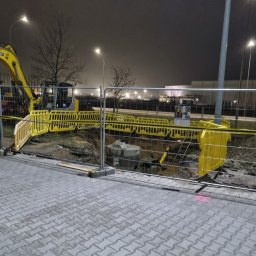 Usługi hydrauliczne wod-kan - Wykop zabezpieczony żółtą barierką, widoczny fragment koparki i betonowy krąg w ziemi, oświetlenie uliczne w tle, nocna sceneria.