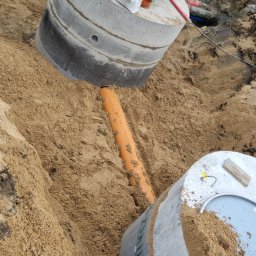 Usługi hydrauliczne wod-kan - Wykonany wykop z widocznymi betonowymi elementami studni kanalizacyjnej i pomarańczową rurą, zasypane piaskiem. Na górnym elemencie studni leżą narzędzia i materiały budowlane.