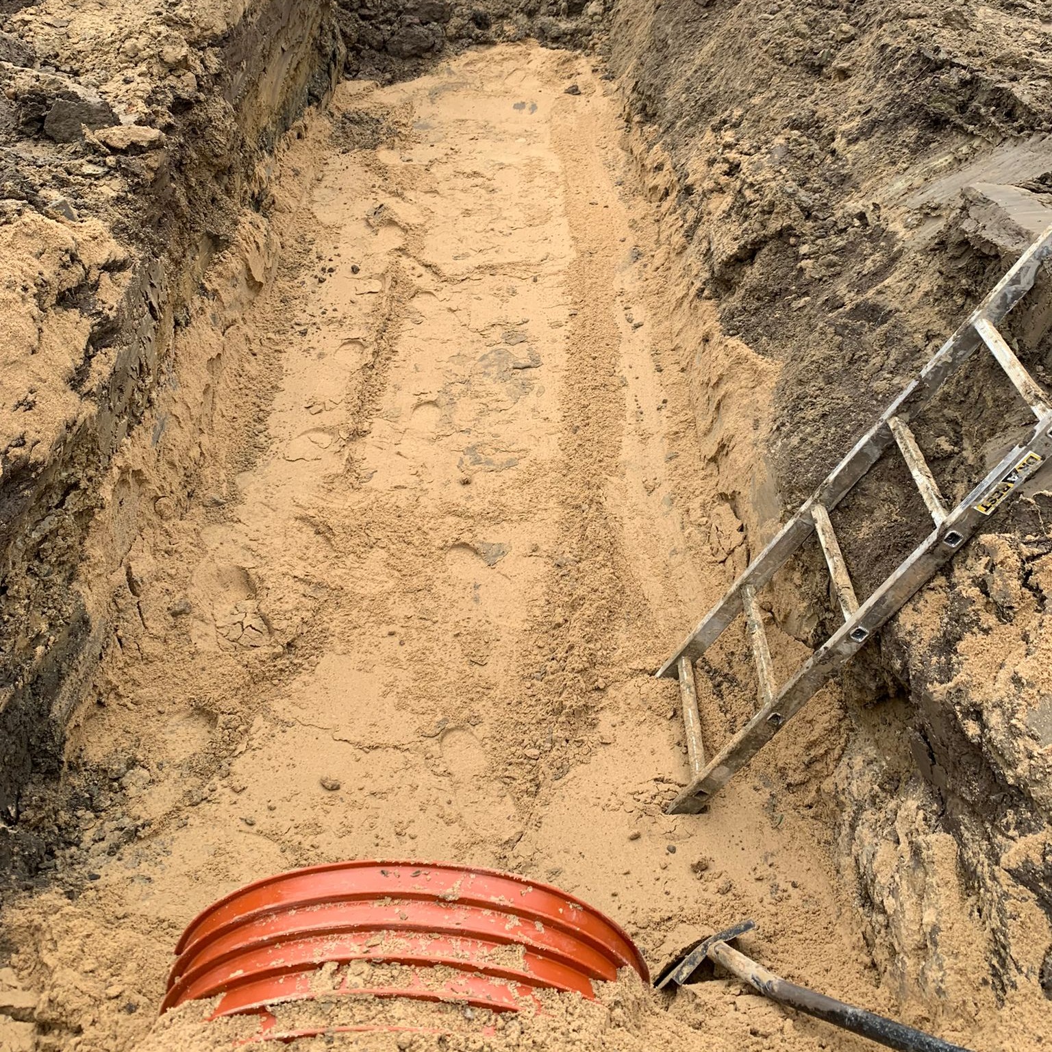 Wykop pod rurę kanalizacyjną z widoczną pomarańczową rurą, aluminiową drabiną opartą o skarpę wykopu i łopatą w piasku.