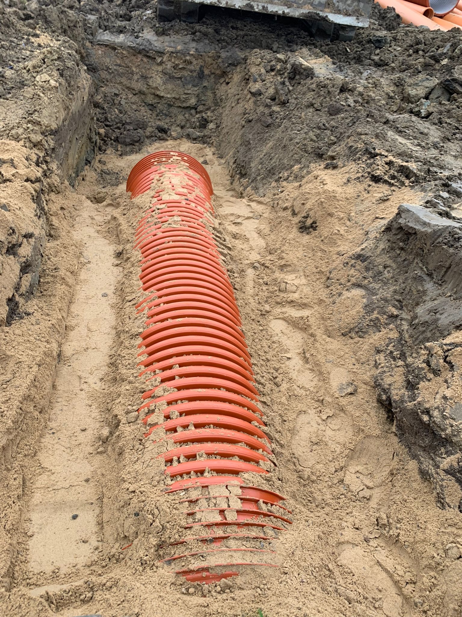 Pomarańczowa, karbowana rura kanalizacyjna ułożona w wykopie, częściowo zasypana piaskiem, z widocznymi śladami stóp na piasku.
