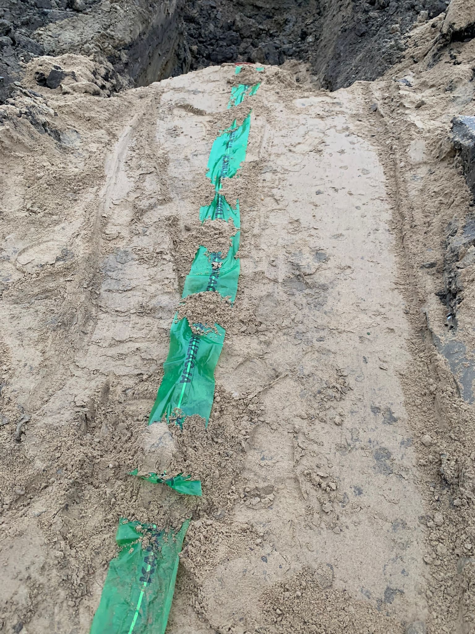 Wykop z ułożonym rurociągiem w zielonej folii ochronnej, widoczne ślady opon i piasek.