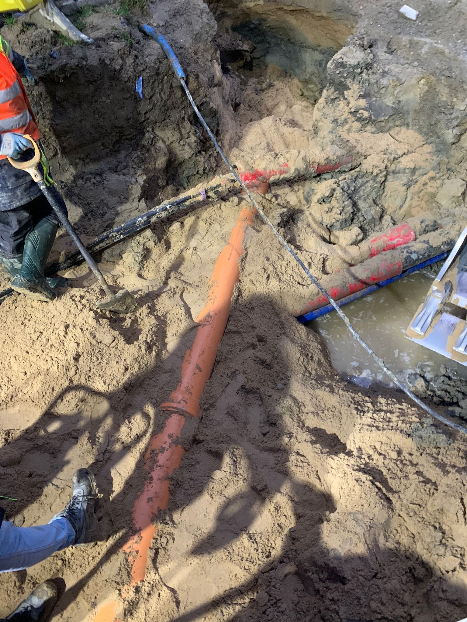 Wykop z widocznymi rurami wodociągowymi i kanalizacyjnymi, częściowo zalany wodą, z pracownikiem w odblaskowej kamizelce i gumowych butach obok łopaty.