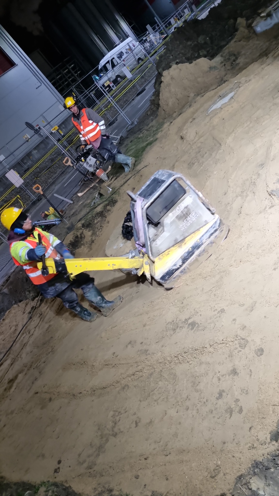 Dwóch pracowników w odblaskowych kamizelkach zagęszcza piasek w wykopie przy użyciu zagęszczarki wibracyjnej HACKER NEUSON, w tle budynek przemysłowy i samochód dostawczy.