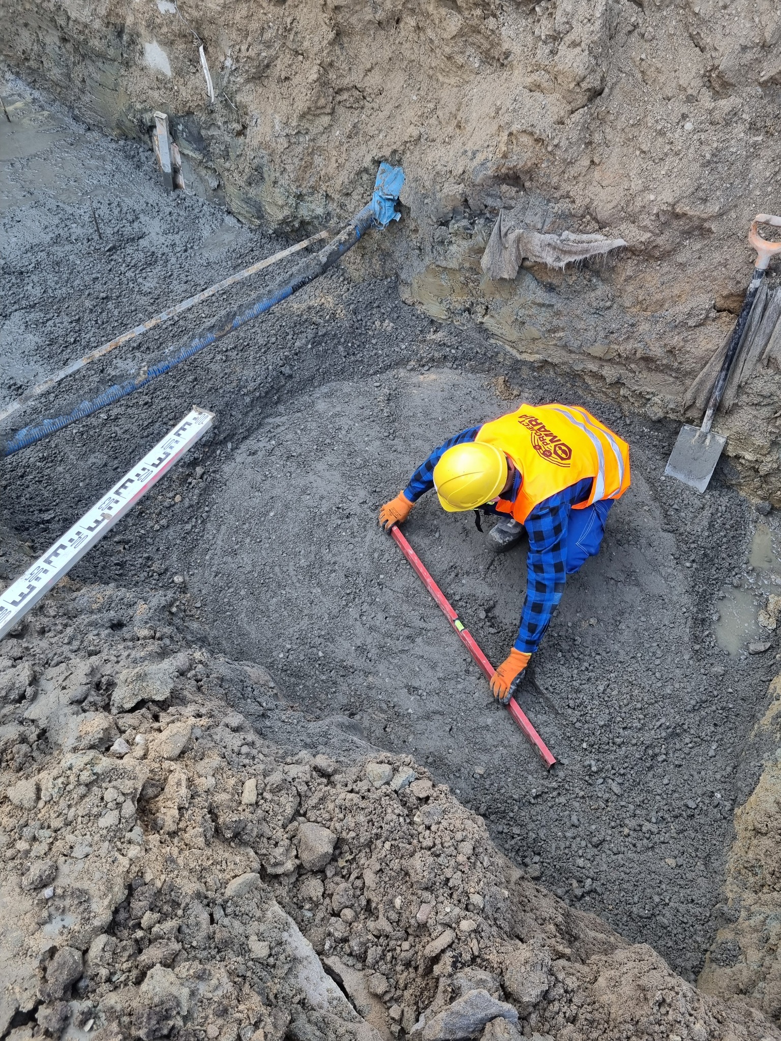 Pracownik w kasku i kamizelce odblaskowej, w wykopie, wyrównuje podłoże poziomnicą. W tle widoczne rury i ściany wykopu.