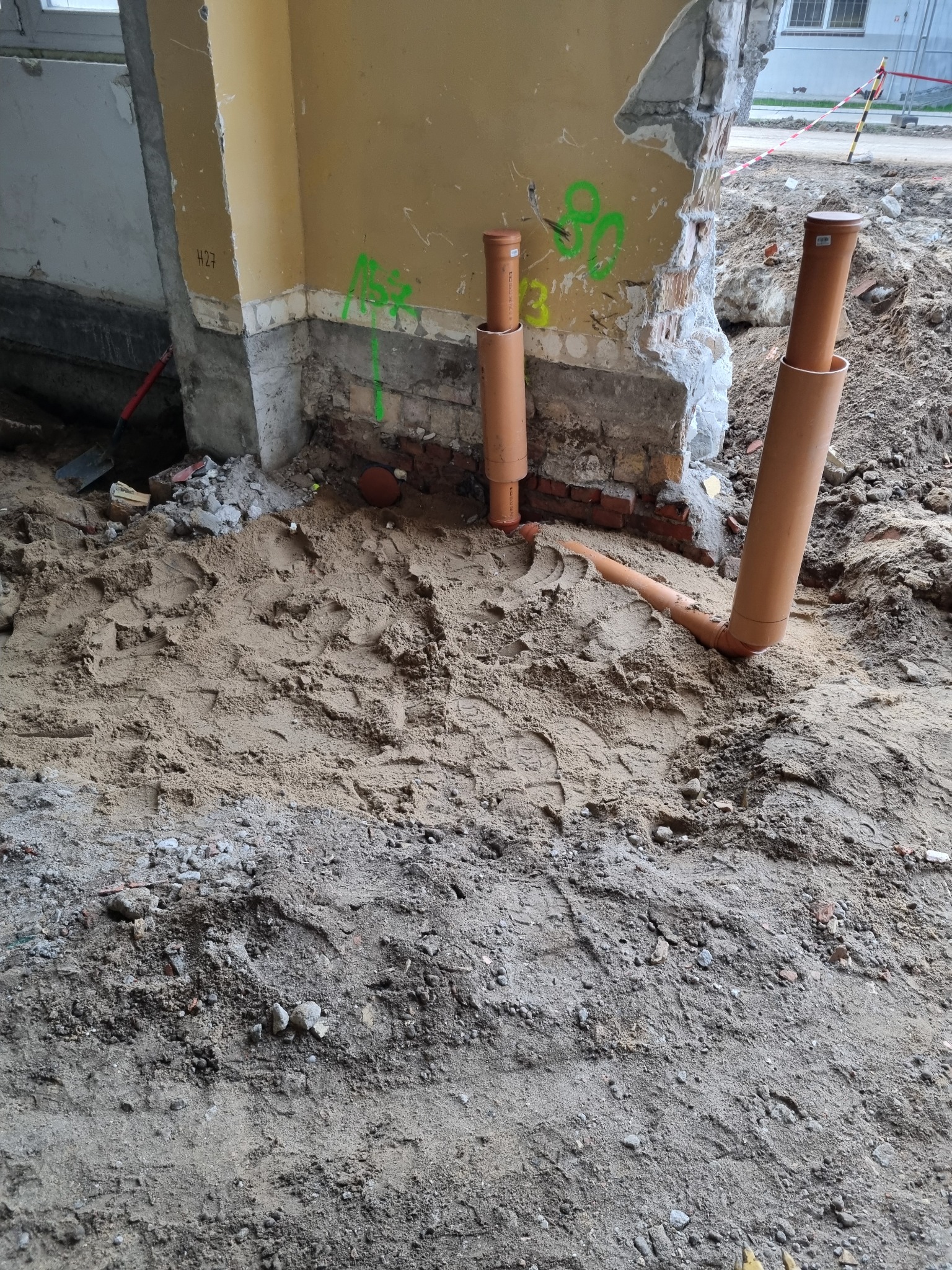 Wykop z piaskiem, fragment ściany z instalacją rur kanalizacyjnych z zaślepkami, ślady zielonych oznaczeń na murze.