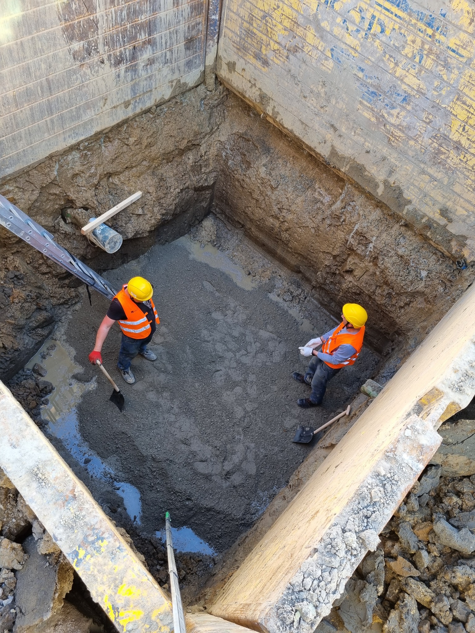 Dwaj pracownicy w kaskach i kamizelkach odblaskowych w wykopie zabezpieczonym szalunkiem, jeden z łopatą, drugi z dokumentami, widok z góry na plac budowy.