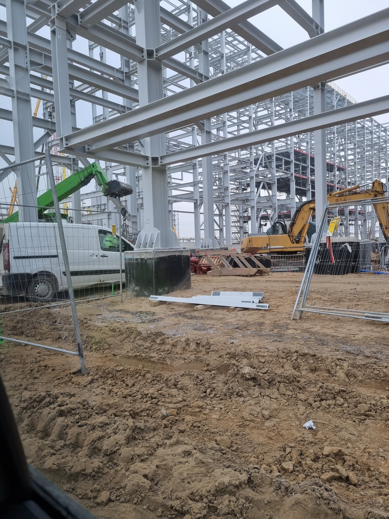 Szkielet stalowej konstrukcji budynku w trakcie montażu, widoczny biały van i koparka, zabezpieczenia budowy w postaci metalowych barierek.
