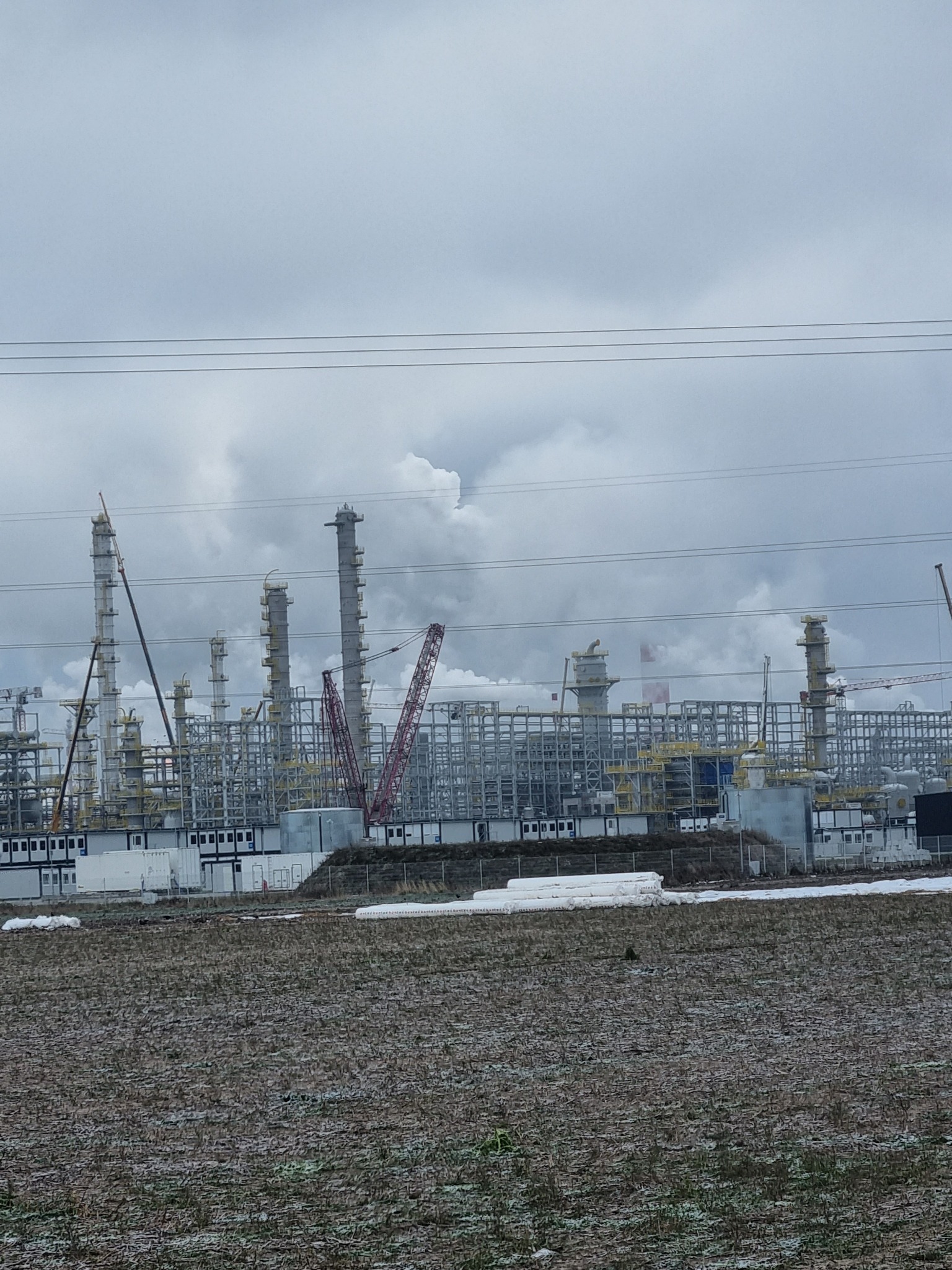 Rozległy widok na budowę instalacji przemysłowej z licznymi kolumnami, rusztowaniami i dźwigami pod pochmurnym niebem, z polem na pierwszym planie.